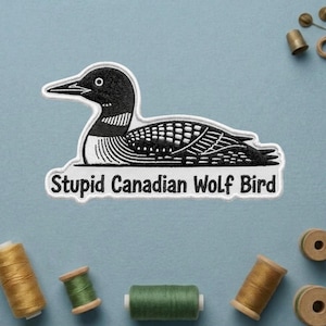 May include: Embroidered patch featuring a stylized black and white loon with the text "Stupid Canadian Wolf Bird". The patch is rectangular with a white border and is likely for clothing or accessories.