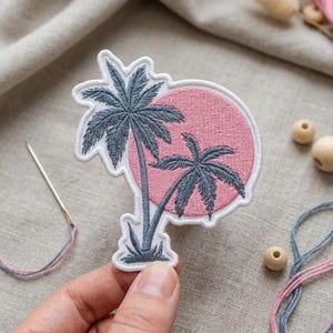 May include: An embroidered patch featuring two dark gray palm trees silhouetted against a solid pink circular sun. The patch has a white border and is shaped to follow the contours of the palm trees and the circular sun design.