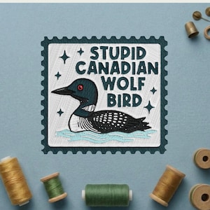 May include: Embroidered patch featuring a loon with the text "STUPID CANADIAN WOLF BIRD" in white. The patch is framed by a dark teal border and four star accents. Sewing supplies, including spools of thread and a thimble, are scattered around the patch.
