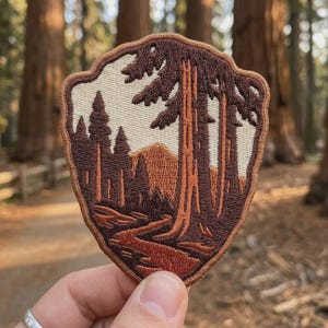 May include: An embroidered patch in a shield shape featuring a landscape scene with tall brown trees, a winding path, and a mountain in the background. The design uses a color palette of cream, dark brown, and light brown threads with a stitched border.