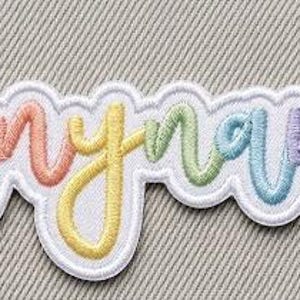 Op de afbeelding: Een gepersonaliseerde geborduurde patch met het woord Anyname in cursief schrift. De letters hebben een regenboogverloop van roze, oranje, gele, groene, blauwe en paarse draad, omlijnd met een dikke witte rand.