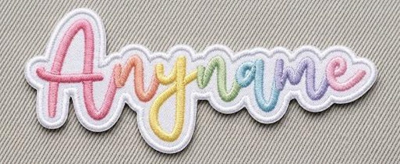 Op de afbeelding: Een gepersonaliseerde geborduurde patch met het woord Anyname in cursief schrift. De letters hebben een regenboogverloop van roze, oranje, gele, groene, blauwe en paarse draad, omlijnd met een dikke witte rand.
