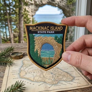 May include: A shield-shaped embroidered patch featuring the text MACKINAC ISLAND STATE PARK in white and gold. The design depicts a tan natural rock archway, green pine trees, and a blue lake. The patch has a gold border and black background.