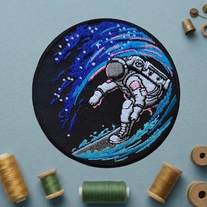 May include: Embroidered patch featuring an astronaut surfing a wave. The astronaut is in a white spacesuit, riding a blue wave against a black circular background. Pink and blue accents create a cosmic effect.
