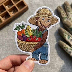 May include: An embroidered patch featuring a brown monkey wearing a tan straw hat and blue denim overalls. The monkey is holding a woven basket filled with colorful vegetables, including orange carrots, purple eggplants, red tomatoes, and corn.