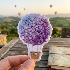 May include: An embroidered patch shaped like a hot air balloon, where the balloon canopy is composed of dense, layered purple and blue hydrangea flower petals. The basket is tan with a woven texture and small decorative pom-pom accents.