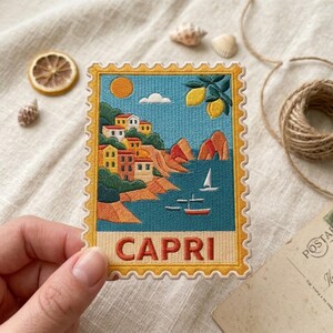May include: A rectangular embroidered patch shaped like a postage stamp featuring a coastal scene of Capri. The design includes colorful houses on a cliff, a bright sun, lemon branches, sailboats on blue water, and the word CAPRI in red letters.