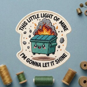 May include: Embroidered patch with the text "THIS LITTLE LIGHT OF MINE I'M GONNA LET IT SHINE". The patch features a teal dumpster on fire with a cute face. The patch is surrounded by a white border.