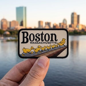 May include: A rectangular embroidered patch with a cream background and a black border. The text reads Boston Massachusetts in black lettering. Below the text, a line of six yellow ducklings walks in a single file line across a brown path.
