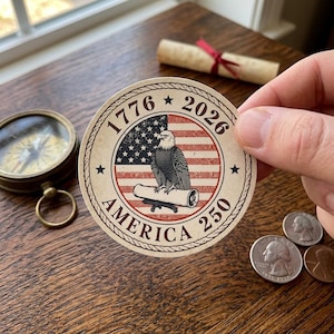 May include: A round sticker with a patriotic design featuring an eagle, the American flag, and the text "1776 2026 AMERICA 250". The sticker is held by a hand, with a compass and coins in the background.