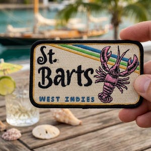 May include: A rectangular embroidered patch with a cream background and a black border. It features the black text St. Barts, blue text West Indies, a pink lobster illustration, and diagonal green and blue stripes across the top right corner.