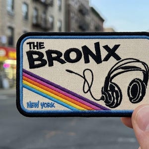 May include: A rectangular embroidered patch with a cream background and a black border. It features the black text THE BRONX and NEW YORK, a black graphic of headphones with a cord, and a diagonal rainbow stripe in red, orange, yellow, and blue.