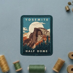 May include: A rectangular embroidered patch featuring a landscape of Yosemite Half Dome with a large white moon in a blue sky. The text YOSEMITE is at the top and HALF DOME is at the bottom in white thread. The patch has a dark teal border.