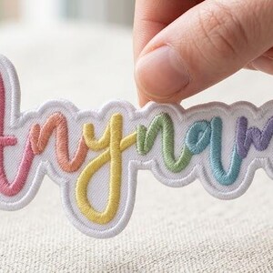 Op de afbeelding: Een op maat gemaakte geborduurde patch met het woord Anyname in een cursief lettertype met een regenboogverloop van roze, oranje, gele, groene, blauwe en paarse draad op een witte achtergrond met een geschulpte witte rand.