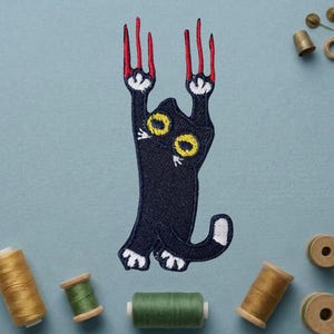 May include: Embroidered patch featuring a black cat with yellow eyes and red claws, in a playful, climbing pose. The cat's paws and tail tip are white. The patch is surrounded by spools of thread in various colors, suggesting a crafting theme.