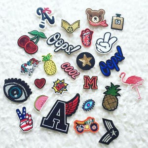 May include: A collection of assorted embroidered iron-on patches featuring various designs including a unicorn, a flamingo, cherries, pineapples, a bear, a motorcycle, eyes, stars, peace signs, the letter A, the letter M, and the word Oops.