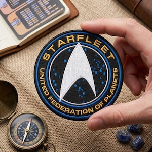 May include: A circular embroidered patch featuring the Starfleet United Federation of Planets emblem. The design includes a white delta symbol on a black background with blue star-like dots, surrounded by a blue border and gold text lettering.