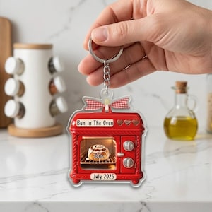 May include: A red oven-shaped keychain featuring a cinnamon roll inside the oven door. The text "Bun in The Oven" is printed at the top, "John" is on the cinnamon roll, and "July 2025" is at the bottom. A red and white checkered bow sits on top.