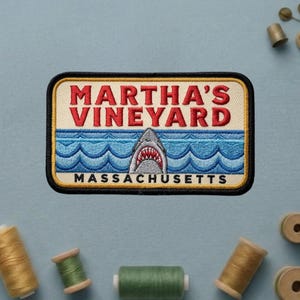 May include: A rectangular embroidered patch featuring the text MARTHA'S VINEYARD in red letters above a graphic of a shark emerging from blue ocean waves, with the word MASSACHUSETTS printed in black at the bottom, all bordered by a black frame.