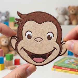 May include: An embroidered patch featuring the face of a cartoon monkey with brown fur, a tan face, large white eyes with brown pupils, and a wide smiling mouth showing a pink tongue. The patch has a dark brown stitched border around the edges.