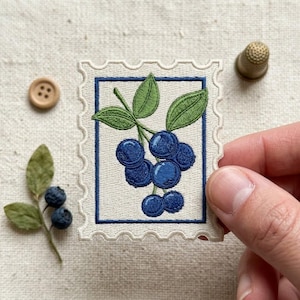 May include: A rectangular embroidered patch shaped like a postage stamp featuring a cluster of dark blue blueberries with green leaves on a cream background, framed by a thin navy blue border, with a small wooden button and thimble nearby.