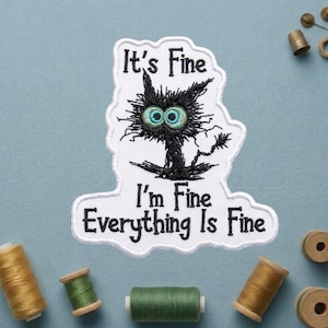 May include: Embroidered patch featuring a black cat with wide, teal eyes and the text "It's Fine, I'm Fine, Everything Is Fine." The patch is white with a black border. Spools of thread in gold and green are scattered around the patch.