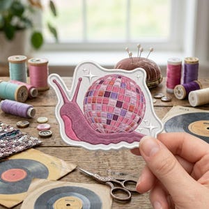 May include: An embroidered patch featuring a pink snail with a disco ball shell in shades of pink and purple, accented with small white star shapes on a white background with a scalloped edge, designed for sewing or decorative application.