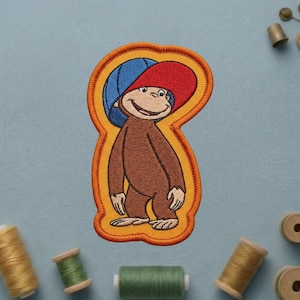 May include: Embroidered patch featuring a cartoon monkey wearing a red hat and a blue hat. The monkey is brown with a smiling face. The patch has an orange border and is on a light blue background.