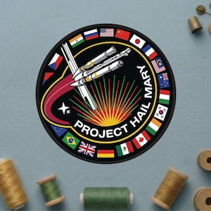 May include: A circular patch with the text "PROJECT HAIL MARY" and a rocket ship design. The patch features a black background with a red and white trail, a sunburst, and flags of various nations around the border. The patch is surrounded by sewing supplies.