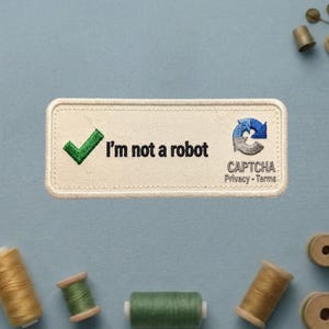 May include: A rectangular embroidered patch with a cream-colored background and a stitched border. It features a green checkmark, the black text "I'm not a robot," and a blue and grey circular arrow icon above the text "CAPTCHA Privacy - Terms."