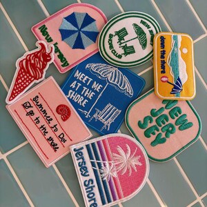 May include: A collection of eight embroidered patches featuring beach-themed designs. The patches include imagery of a red ice cream cone, a blue umbrella, a beach chair, palm trees, and text such as New Jersey, Shore Club, and Summer to Do.