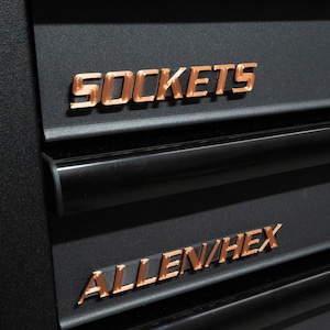 Może przedstawiać: Zbliżenie czarnej szuflady skrzynki narzędziowej z miedzianymi literami. Słowo "SOCKETS" jest wyświetlane nad "ALLEN/HEX". Litery są pogrubione, bezszeryfowe. Szuflada ma czarny uchwyt.