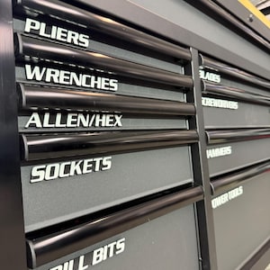 May include: A black metal tool chest with multiple drawers. Each drawer is labeled with white text indicating the contents: PLIERS, WRENCHES, ALLEN/HEX, SOCKETS, DRILL BITS, BLADES, SCREWDRIVERS, HAMMERS, and POWER TOOLS. The chest has black handles.