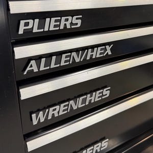 May include: Close-up of a black tool chest with silver labels. The labels read: SCREWDRIVERS, PLIERS, ALLEN/HEX, WRENCHES, and HAMMERS. The chest has horizontal silver stripes between the drawers.