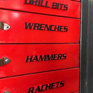 Puede incluir: Un cofre de herramientas rojo con letras negras. Los cajones están etiquetados como "DRILL BITS", "WRENCHES", "HAMMERS" y "RACHETS". Cada cajón tiene un asa plateada.