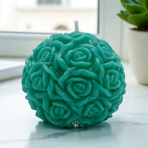 May include: A teal-colored, spherical candle with a detailed rose design. The candle has a single white wick and is placed on a white surface. The candle's surface is covered in sculpted roses, creating a textured appearance.