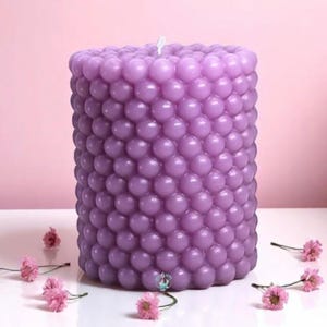 May include: A large, lavender-colored pillar candle with a textured, bubble-like surface. The candle is cylindrical and stands upright on a white surface, with small pink flowers scattered around its base. The background is a soft pink.