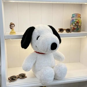 May include: A plush Snoopy dog toy with black ears, a black nose, and a red collar. The white stuffed animal is sitting on a shelf with other decorative items, including sunglasses and a jar filled with colorful characters.