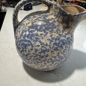 Puede incluir: Una jarra de cerámica redonda con un patrón de esponjado o moteado en azul y crema. El recipiente tiene un asa curva en el lateral y un pico vertedor ancho y abierto en la parte superior, con un acabado liso y brillante en toda la superficie.