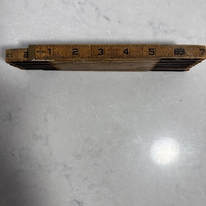 May include: A folded wooden ruler with a natural tan finish and black markings. The ruler features clear numerical increments in inches, with a metal hinge visible on the right side and a metal end cap on the left, showing measurements up to 7 inches.