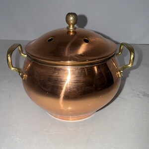 Puede incluir: Una olla de cobre pulido con cuerpo redondeado, que cuenta con dos asas laterales de latón y una tapa abovedada a juego. La tapa está rematada con un pequeño pomo de latón e incluye dos pequeños orificios de ventilación circulares.