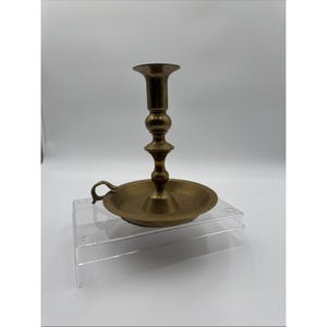 Puede incluir: Un candelabro de latón con una base circular ancha que sirve como bandeja para gotas de cera y un asa pequeña y curva en el lateral. El vástago central presenta un diseño torneado de varios niveles con una abertura superior ensanchada.