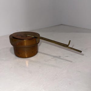 Op de afbeelding: Een cilindervormig, bronskleurig metalen gewicht met een platte bovenkant en een lange, dunne, bevestigde metalen arm die horizontaal uitsteekt. Het bovenoppervlak is gegraveerd met het getal 100 en de afwerking vertoont sporen van slijtage en patina.