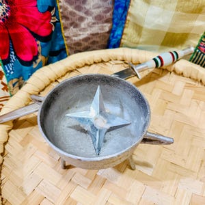 Puede incluir: Un cuenco de metal plateado con un diseño en forma de estrella en el centro, apoyado sobre una bandeja tejida. Un pequeño cuchillo con mango blanco, verde y rojo está colocado en la bandeja. El cuenco tiene tres patas pequeñas y dos asas.