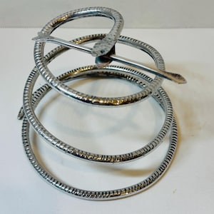 May include: A silver-toned metal spiral stand featuring a textured surface and a snake-shaped topper. The object consists of three graduated circular rings connected in a coil formation, with the snake head and tail crossing at the top point.