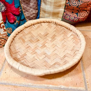 May include: A round, woven bamboo tray with a light tan color. The tray has a tightly woven pattern and a wrapped edge. The tray is sitting on a light brown tile surface. Decorative fabric pillows are in the background.