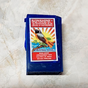 May include: A dark blue box with a label featuring a bird illustration and the text "SUNSHINE ULTRA MARINE BLUE". The label also includes the text "ORIGINAL BLUE" and the manufacturer's details.