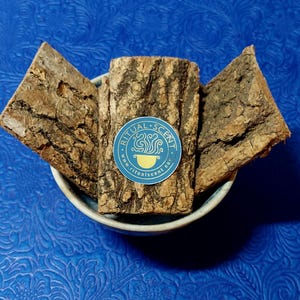 Puede incluir: Un cuenco azul claro contiene tres trozos de corteza de árbol con una superficie marrón texturizada. Una pegatina azul con el texto "RITUAL SCENT" y un logotipo está adherida a la corteza. El fondo es una superficie azul texturizada.