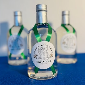 Könnte beinhalten: Drei klare Glasflaschen Siwo Doja Sirup, jede mit einem silbernen Verschluss und einem grünen Band. Die Flaschen haben ein weißes Etikett mit Text und einem dekorativen Design. Die Flaschen stehen auf einer blauen Oberfläche.