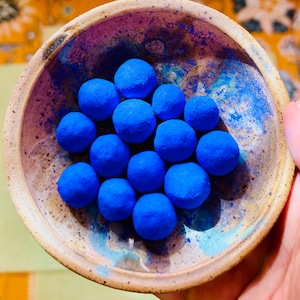 May include: A ceramic bowl with a speckled, earthy tone holds a collection of vibrant blue spheres. The bowl's interior features a splash of blue hues, complementing the spherical objects. The arrangement creates a visually appealing contrast of textures and colors.